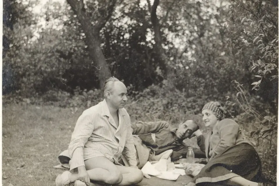 Photographie ancienne en noir et blanc : trois personnes pique-niquent sur l'herbe à l'ombre des arbres